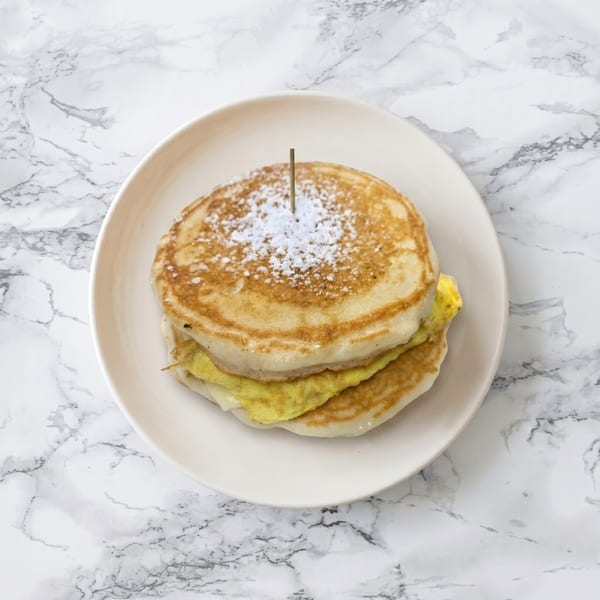 A stack of pancakes with scrambled eggs and cheese, topped with powdered sugar and a toothpick.